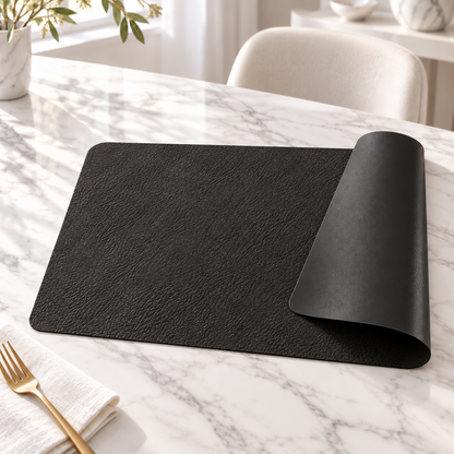 Black reversible faux leather placemat on a white marble table with the corner curled to show the smooth back